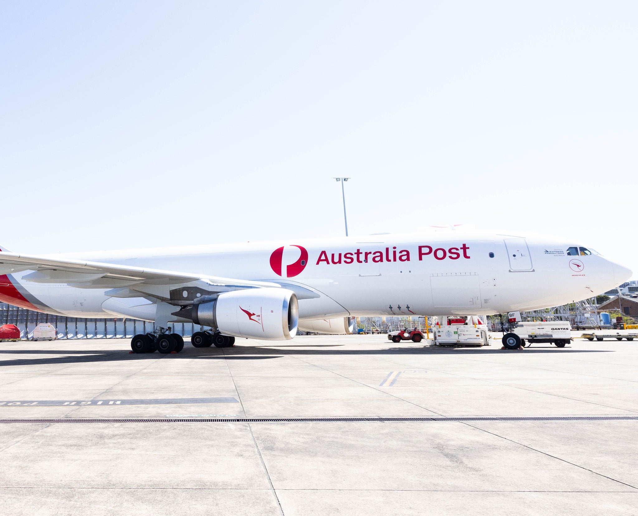 A large white cargo airplane with Australia Post markings is parked on an airport tarmac. The aircraft is viewed from the side, showing its fuselage, wings, and engines in bright daylight. Ground support vehicles and equipment are visible near the plane, indicating loading or unloading operations. The clear sky and open apron emphasize an active logistics and air freight environment.