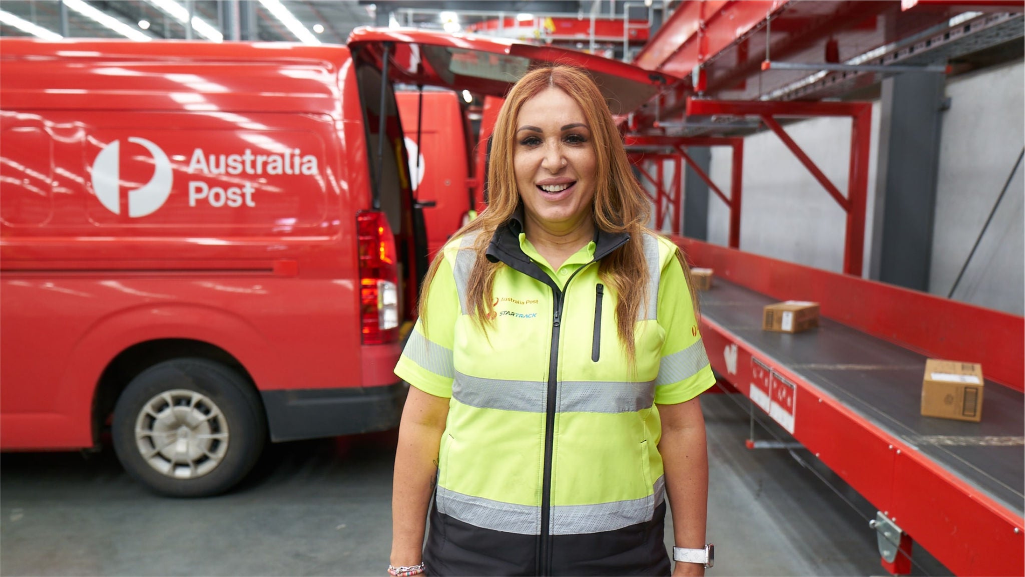 A woman in an Australia POst usiform standing in front of a red Post van loading/unloading parcels to a conveyor belt