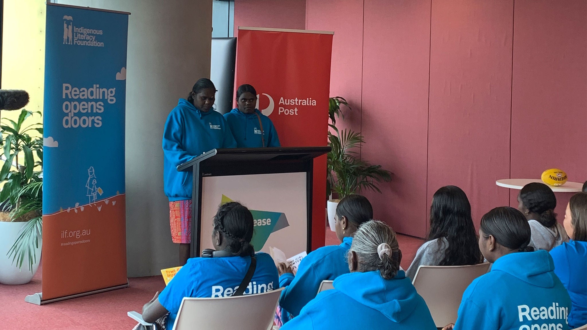 photo-two-first-nations-girls-presenting-in-front-of-a-group-standing-in-front-of-indigenous-literacy-foundation-and-australia-post-banners
