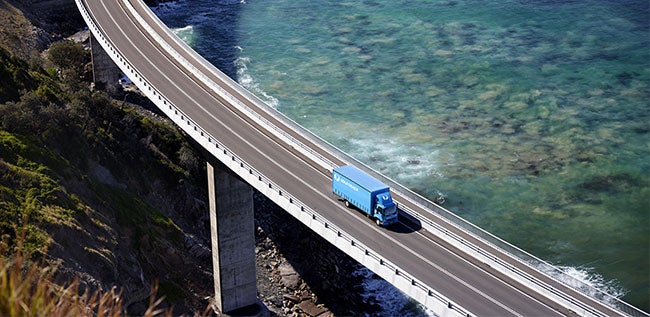 aerial-view-of-a-blue-startrack-truck-driving-along-a-highway-with-the-ocean-on-one-side