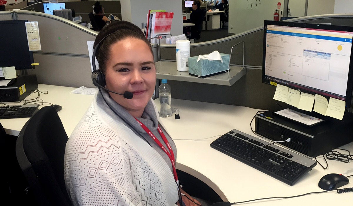 A woman sitting at a call centre desk in from of a computer screen wearing a headset with a microphone